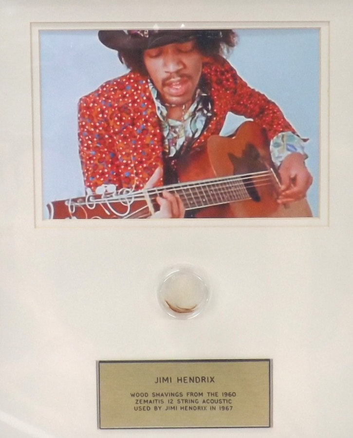 Lot Number 661. Jimi Hendrix - wood shavings from the 1960 Zemaitis acoustic guitar played by Jimi Hendrix, framed between a plaque and photograph of Jimi playing the guitar *From the collection of Keith Smart, sold with a letter of provenance from Keith stating 'I run the Zemaitis Guitar Owners Club.  A luthier friend of mine (and Z Club member) worked on the Hendrix Zemaitis acoustic, knowing my penchant for memorabilia, he sent me some shavings.  I donated some to the Boat Project who made a sailing boat out of 1000 donated bits of wood.  The Hendrix shavings are in a capsule in the tiller.  It was featured in the newspapers and on various television news reports.  I have included copies of magazines featuring articles'. Auctioned at Memorabilia, Guitar Amps, Effects & Audio on 13th June 2019